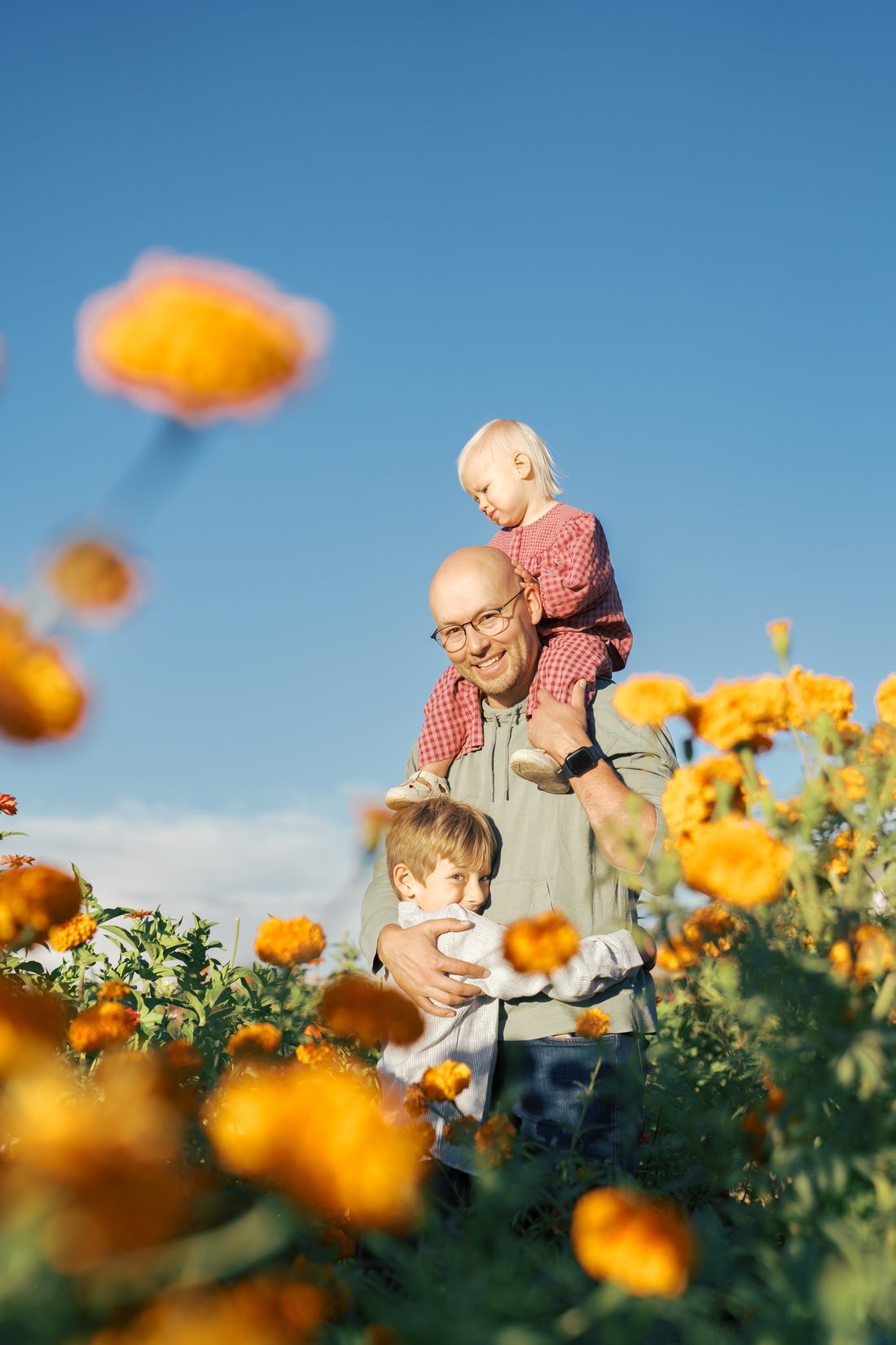 Dad with daughter on his shoulders and son hugging him in a marigold field