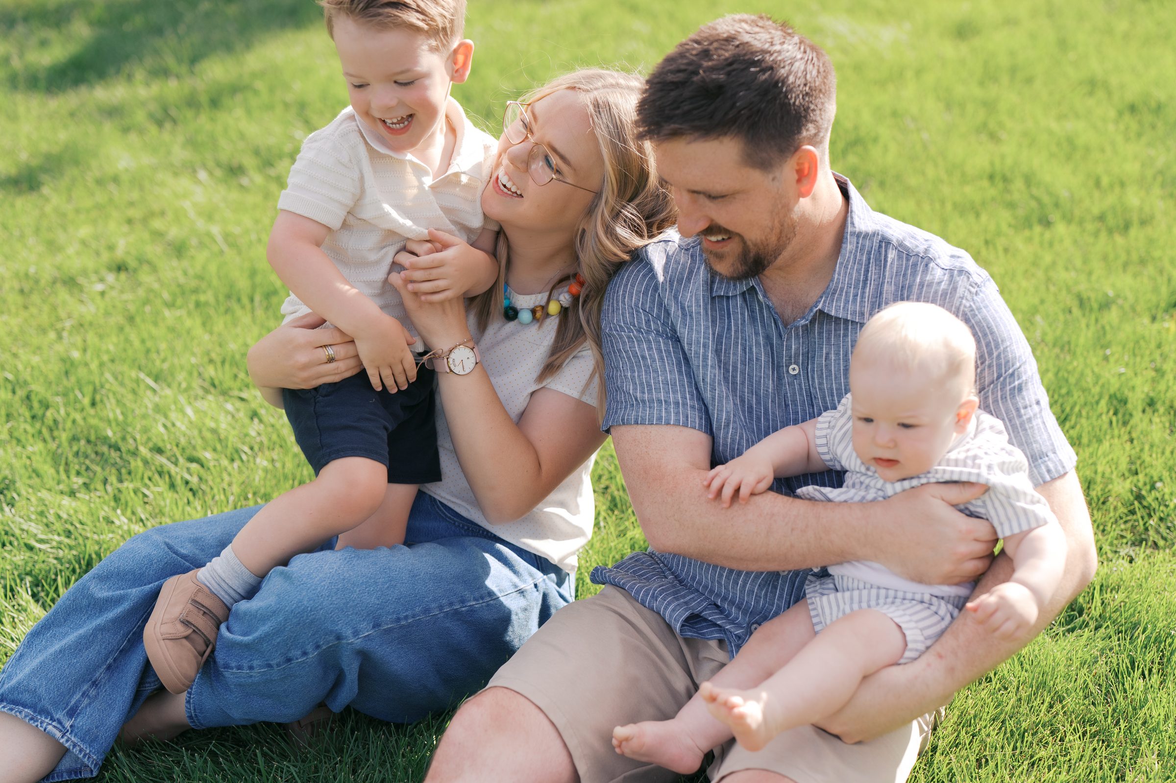 Utah family laughing together on the grass at golden hour, mom holding toddler and dad holding baby