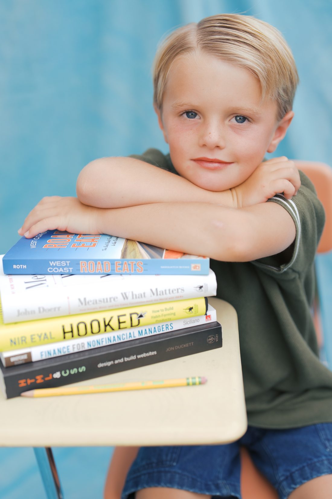 Boy resting his arms on a stack of schoolbooks at a vintage desk, smiling softly at the camera