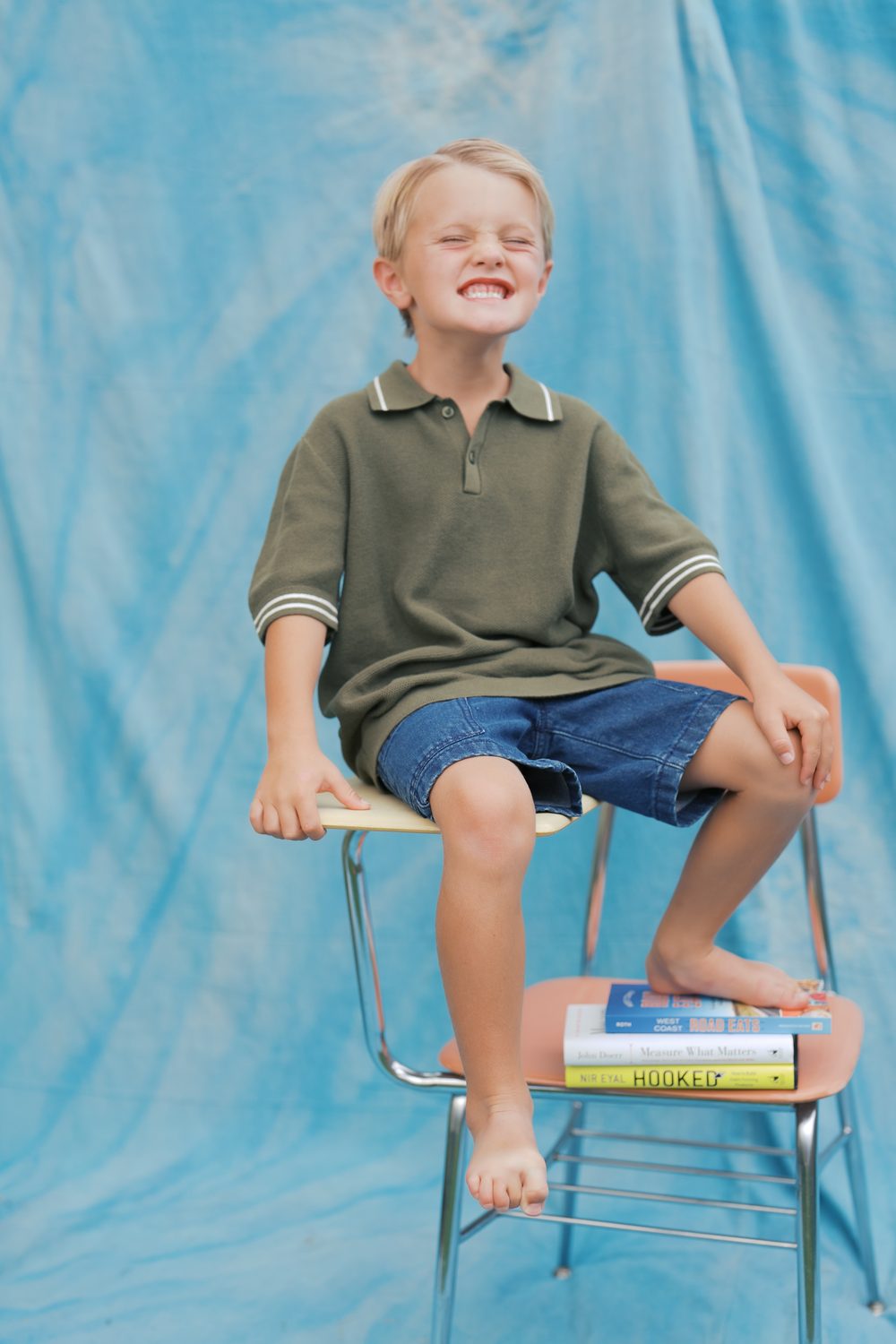 Boy laughing with his eyes squeezed shut, perched barefoot on a retro school desk with his feet on a stack of books
