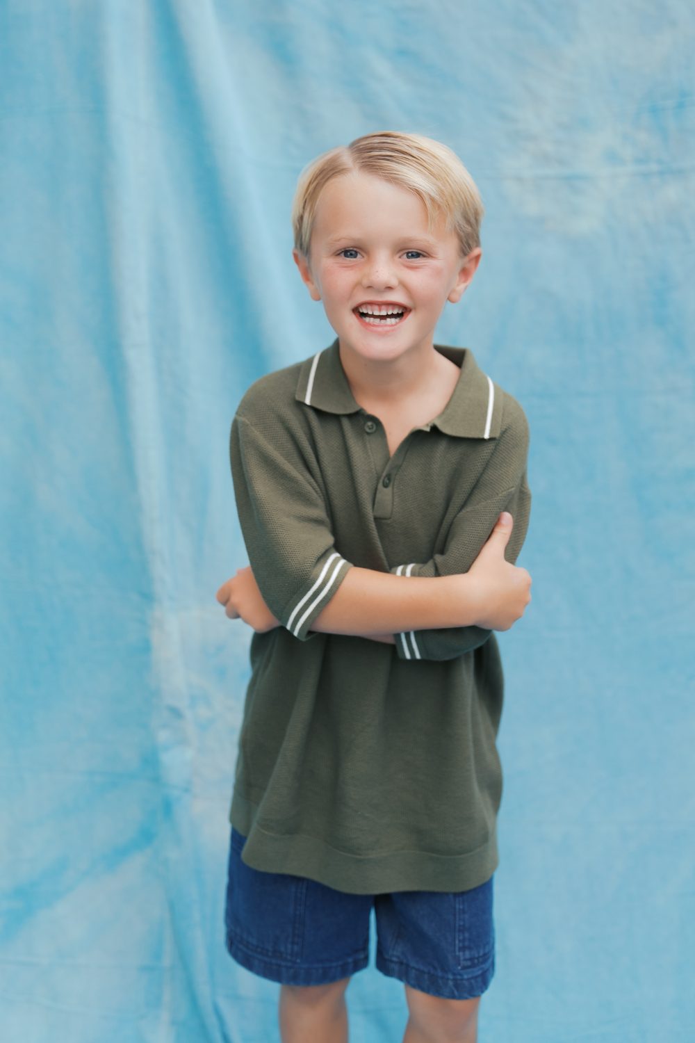 Boy in an olive polo with his arms crossed, laughing at the camera against a hand-dyed blue backdrop