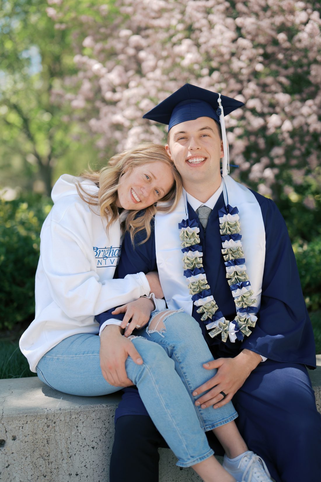 BYU graduate in cap and gown with his wife resting her head on his shoulder under spring blossoms