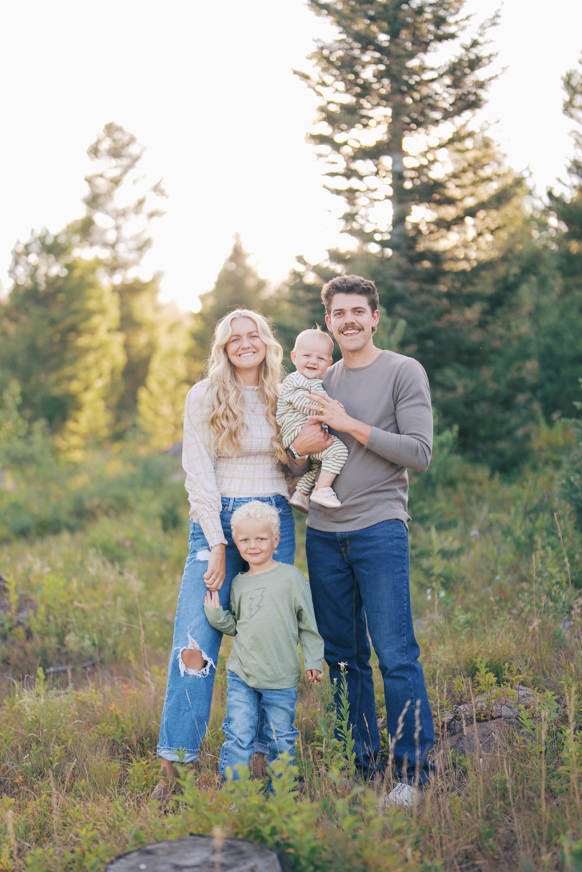 Blonde family of four with curly-haired kids smiling in a golden pine meadow