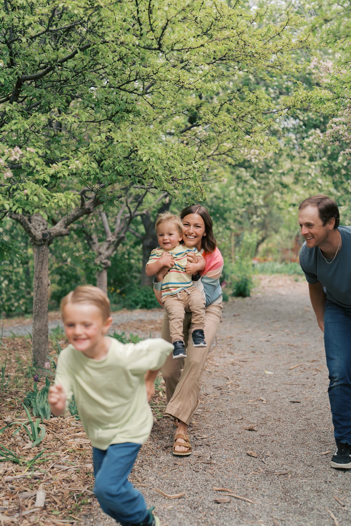 Boy racing down an orchard path while mom laughs holding his little brother