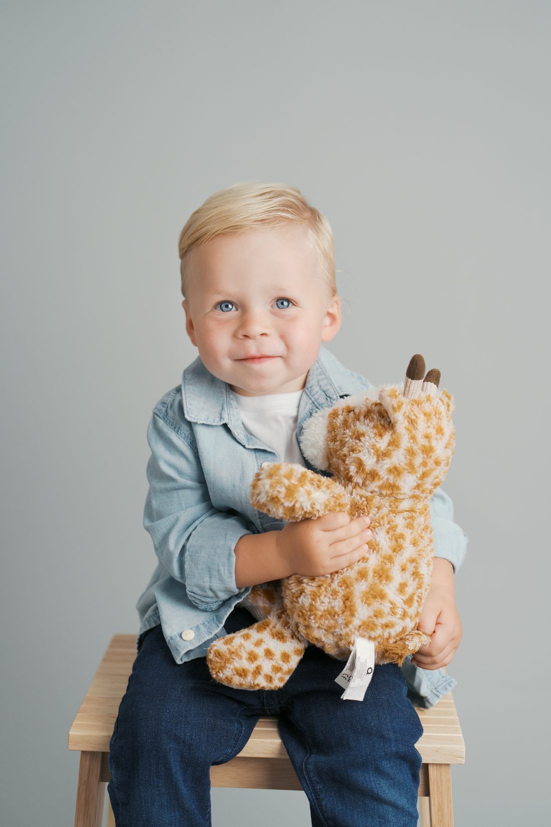 Toddler in a denim shirt hugging his stuffed giraffe on a studio stool