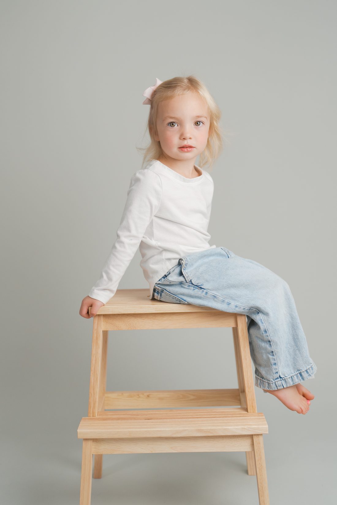 Little girl with a pink bow perched on a wooden stool in the studio