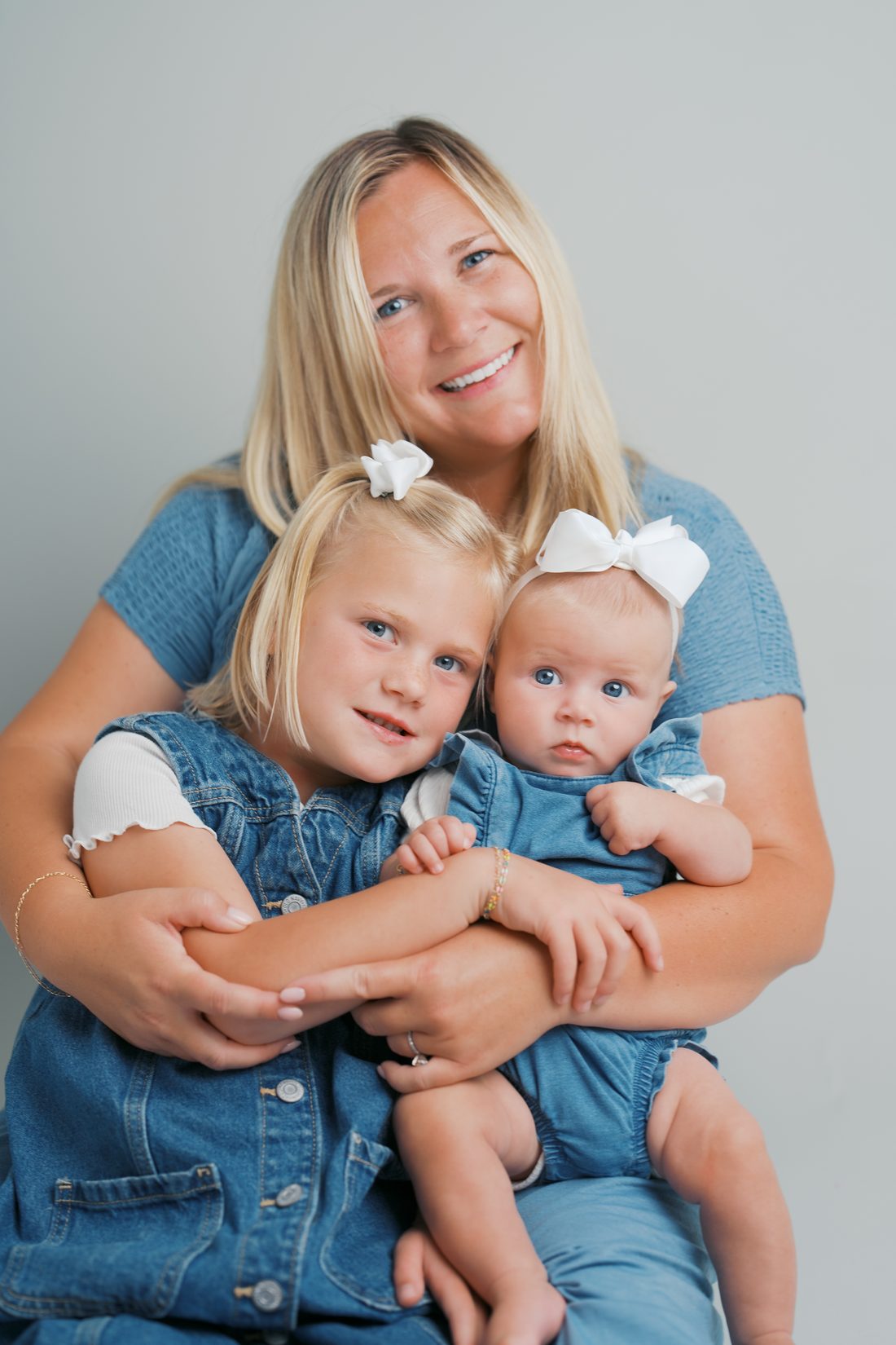 Mom in denim cuddling her two daughters in matching white bows in the studio