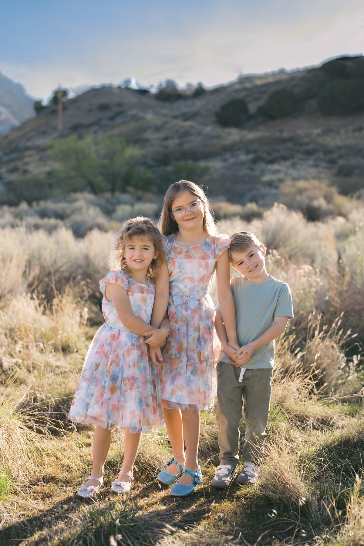 Three siblings in floral dresses and sage green standing on a golden Utah hillside
