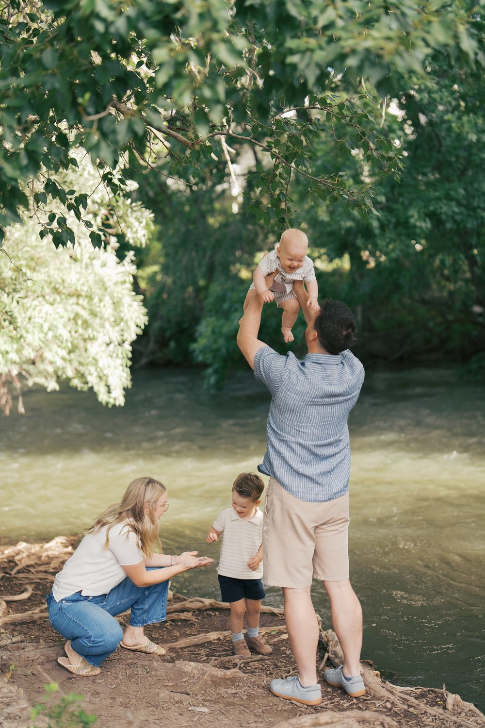 Dad lifting laughing baby into the air by a creek while mom and brother play